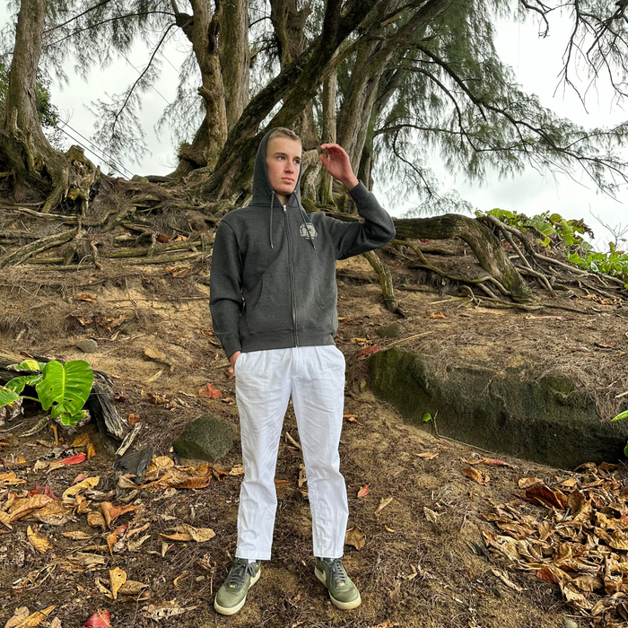 Person wearing a Dog Beach Zip hoodie and white pants standing on a sandy forest floor with trees and fallen leaves in the background.